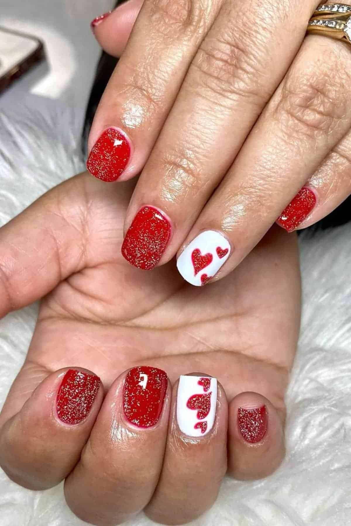 A hand with red glitter nail polish; two nails have white polish with red heart designs.