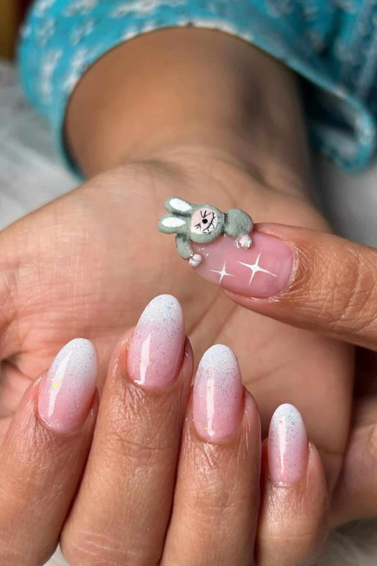 A hand with pink ombre almond-shaped nails; the thumb nail features a 3D design of a small gray and white bunny with a pink face.