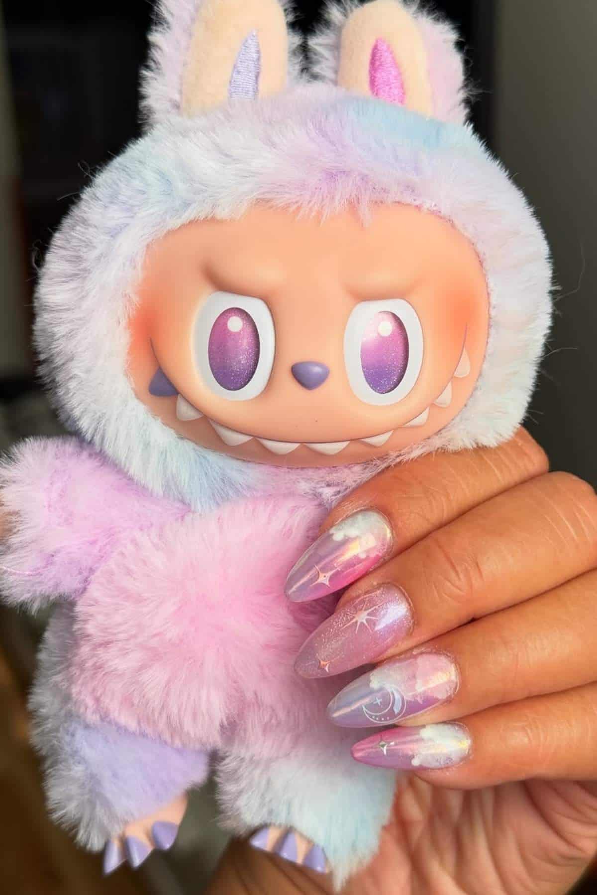 A hand with long, pastel-themed, cosmic nail art holds a plush toy in a pastel, bunny-themed suit with wide eyes and pointed teeth.