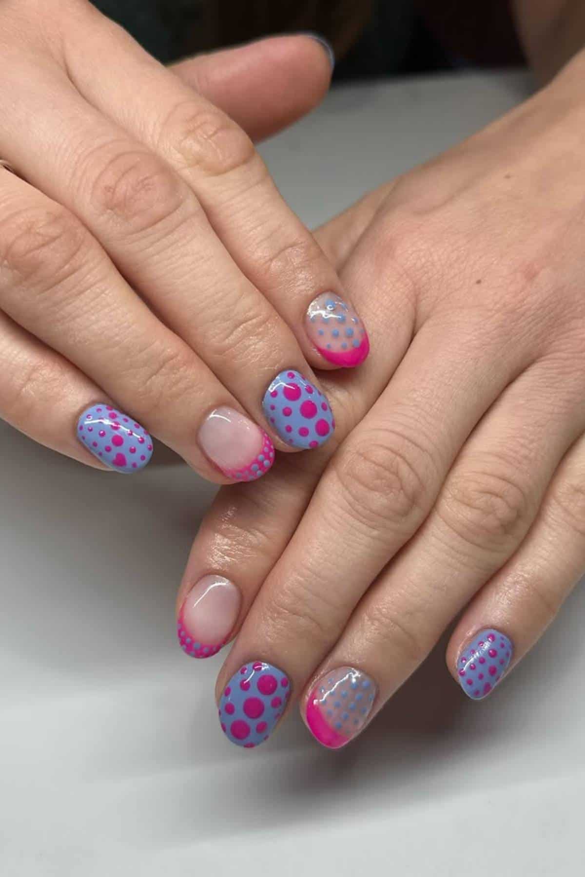 Hands with manicured nails painted in pastel blue and pink with polka dot patterns and pink tips on some nails, shown resting on a white surface.