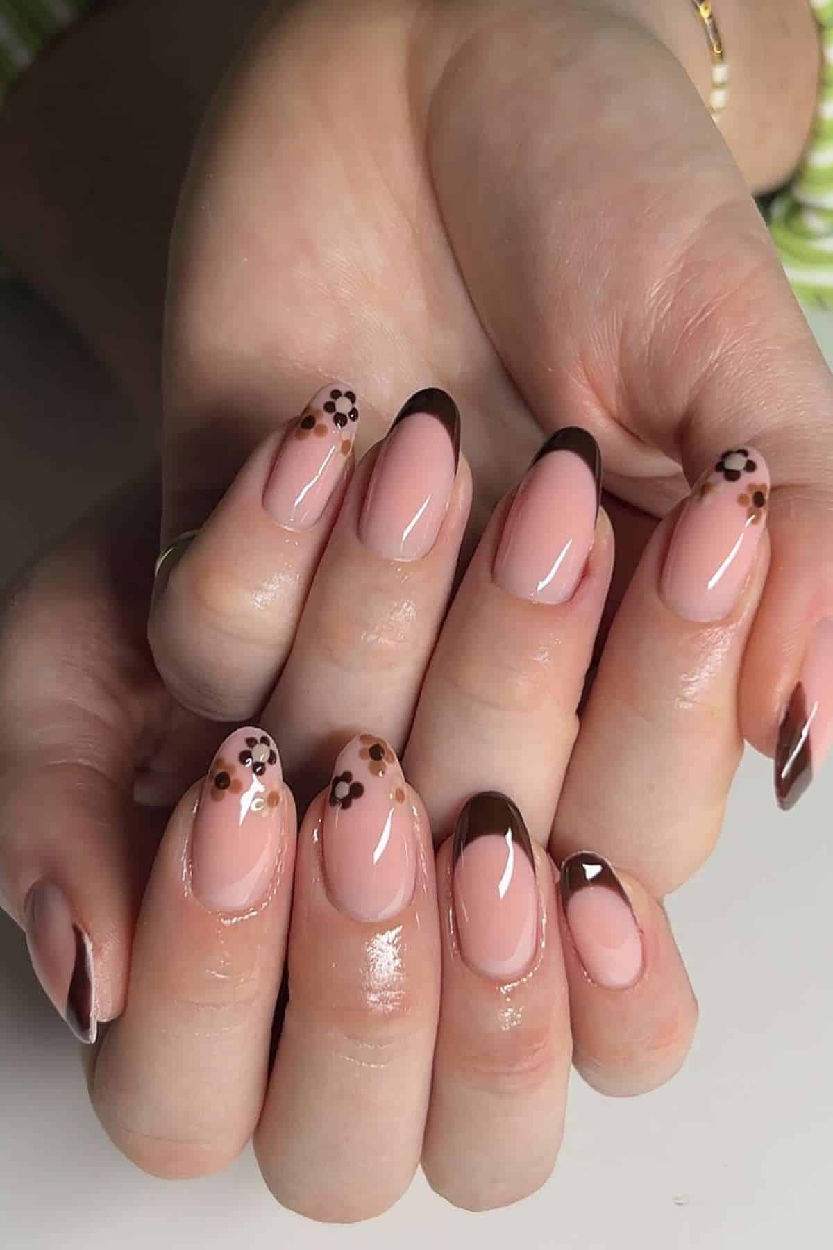Hands with manicured nails featuring glossy nude polish, brown French tips, and small brown and black flower designs.