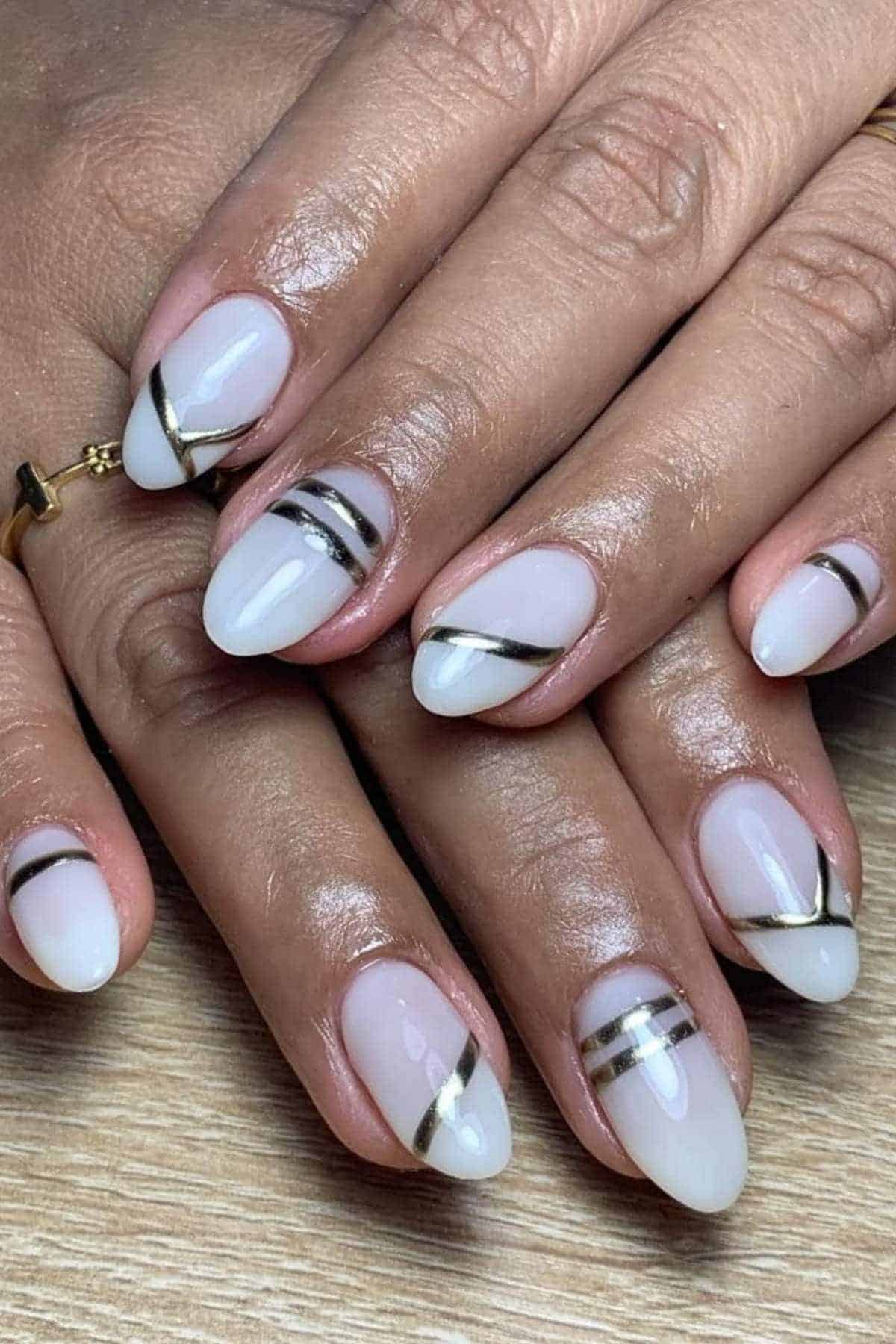 Hands with almond-shaped nails painted in a sheer nude color, featuring black and gold metallic diagonal stripe designs on each nail.