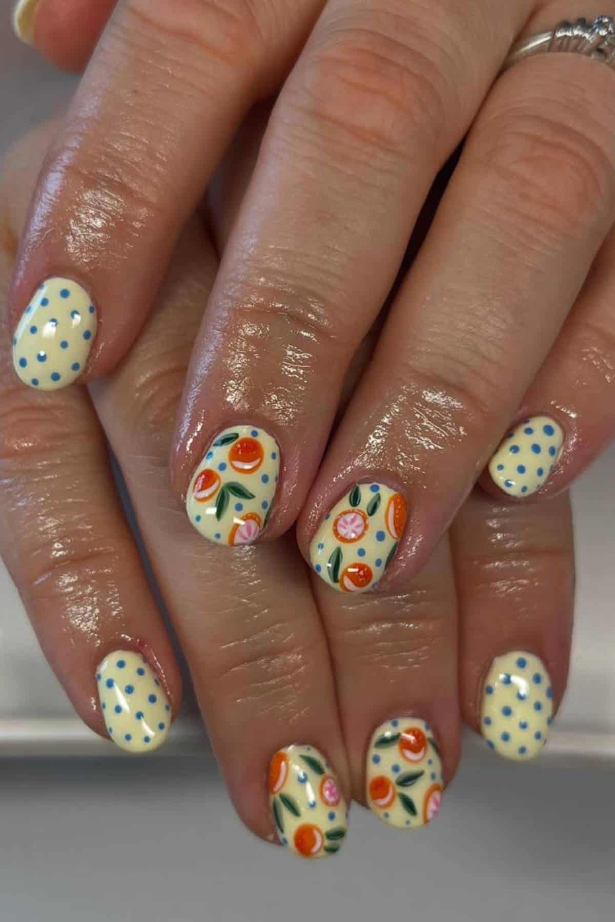Hands with a yellow manicure featuring blue polka dots on some nails and colorful orange, pink, and green fruit patterns on others.