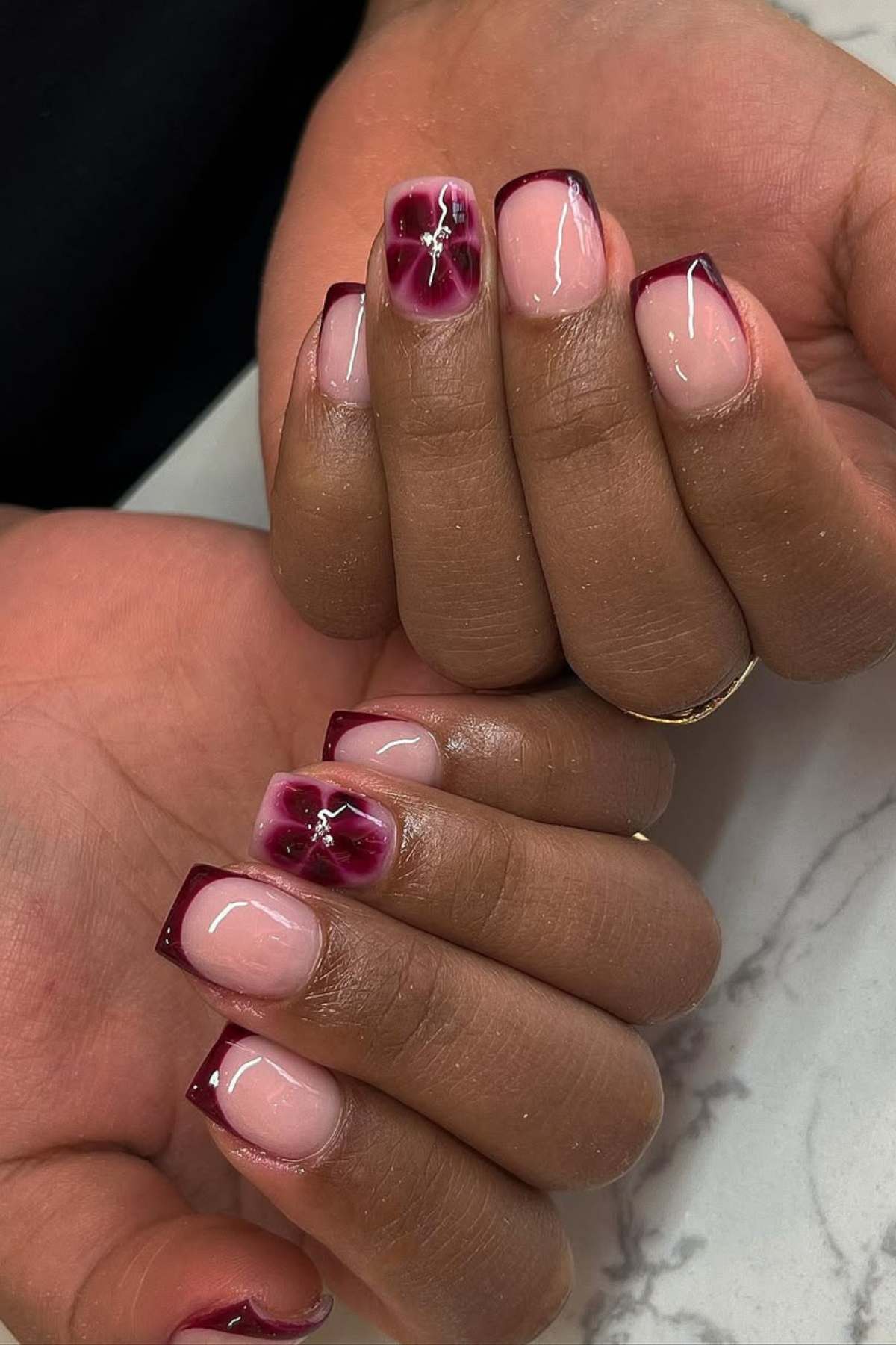 Hands with square-shaped nails featuring a pink base, dark burgundy French tips, and accent nails with a dark burgundy flower design and small rhinestone detail.