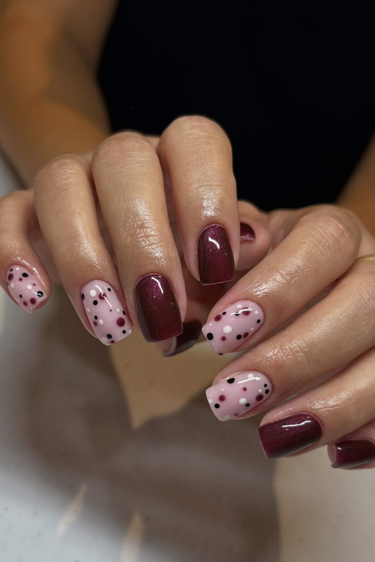 A hand with manicured nails, featuring alternating dark burgundy polish and light pink polish with black and burgundy polka dot designs.