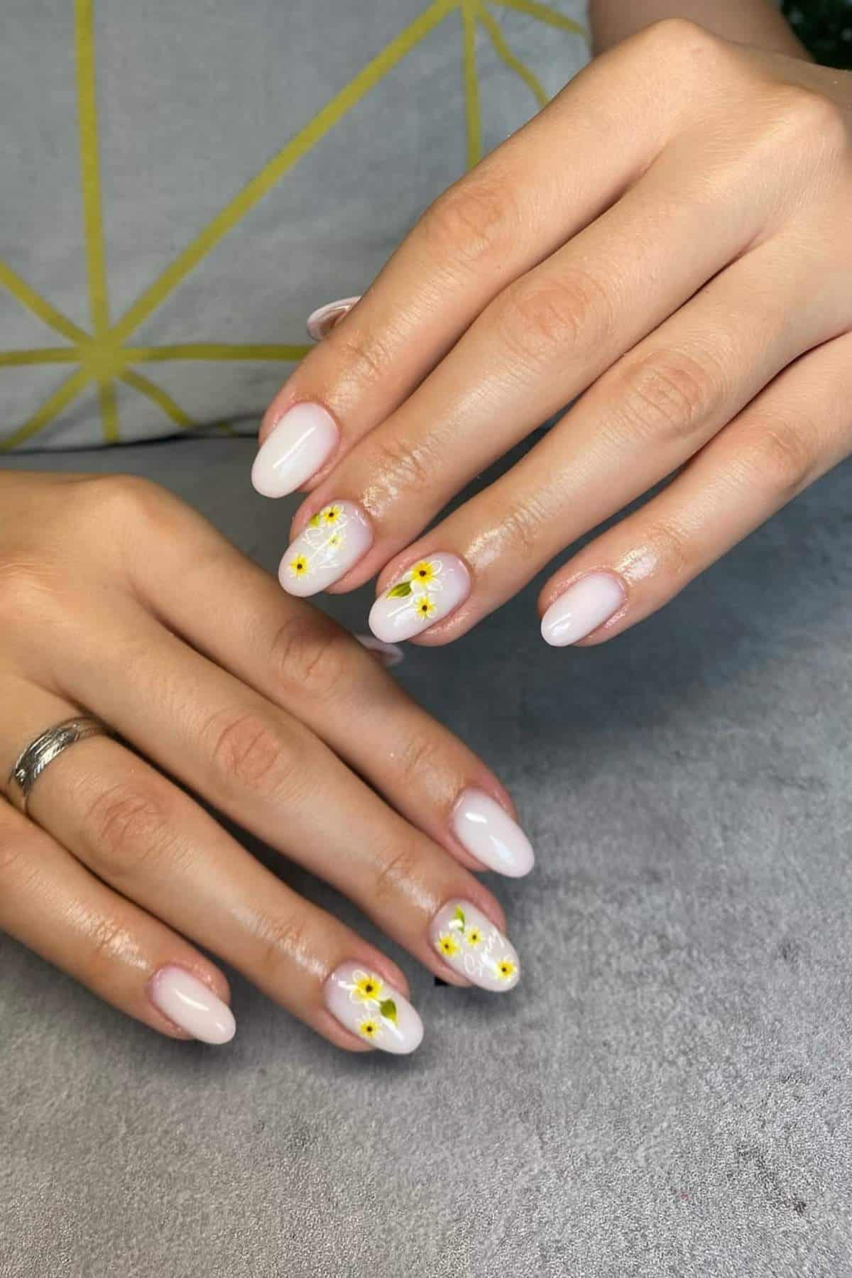 Hands with almond-shaped, pale pink nails; ring finger and middle finger feature small yellow floral designs with green accents on each hand.