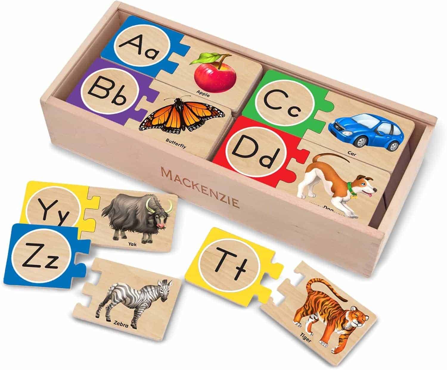 Wooden alphabet puzzle pieces with letters, matching words, and illustrations of objects or animals, arranged inside and outside a wooden box labeled “MACKENZIE.”.