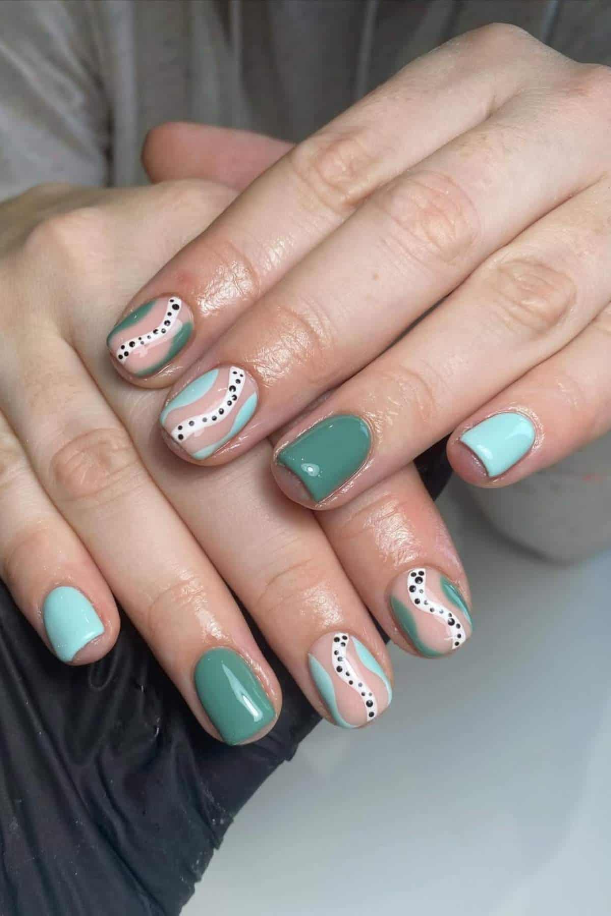 Hands with short nails painted in mint green and light blue, featuring abstract white, beige, and black dotted line designs on several fingers.