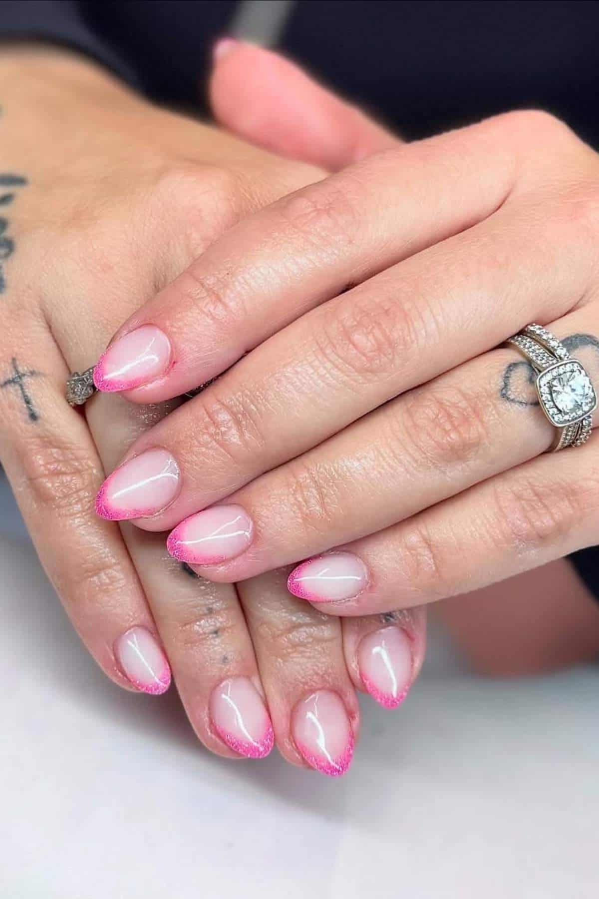 Close-up of hands with pink ombre French manicure, almond-shaped nails, tattoos on fingers, and a diamond engagement ring on the ring finger.