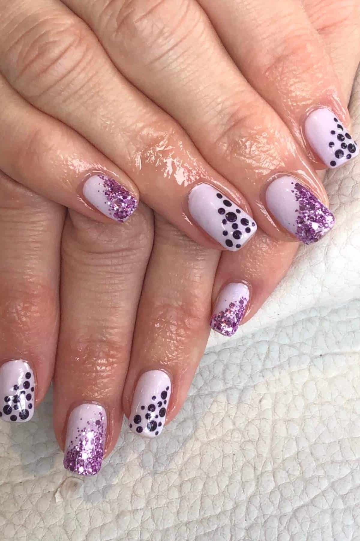 Hands with light purple nail polish featuring purple glitter and black polka dot designs on short, square-shaped fingernails.