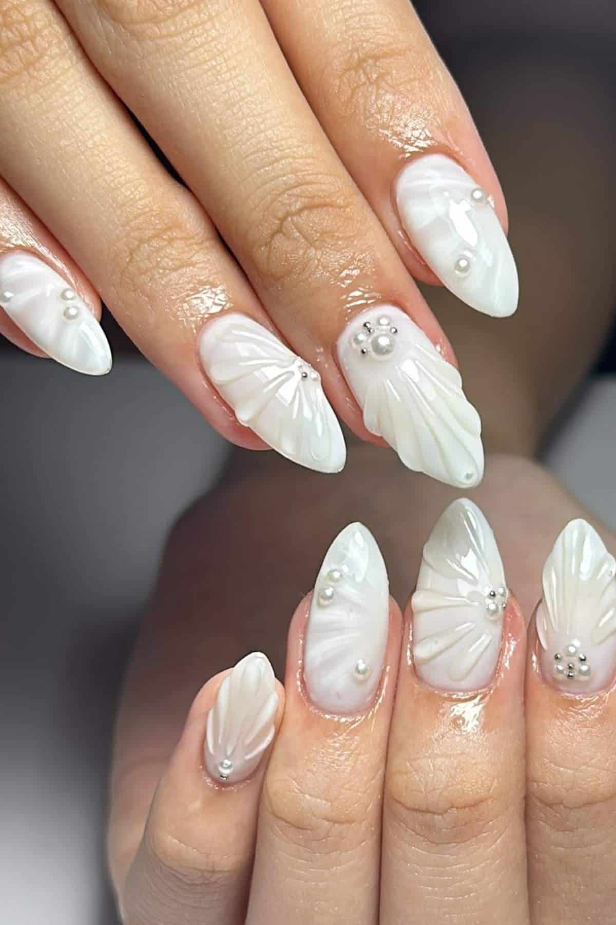 Close-up of hands with almond-shaped nails featuring a white, 3D floral design and small pearl accents on a glossy finish.