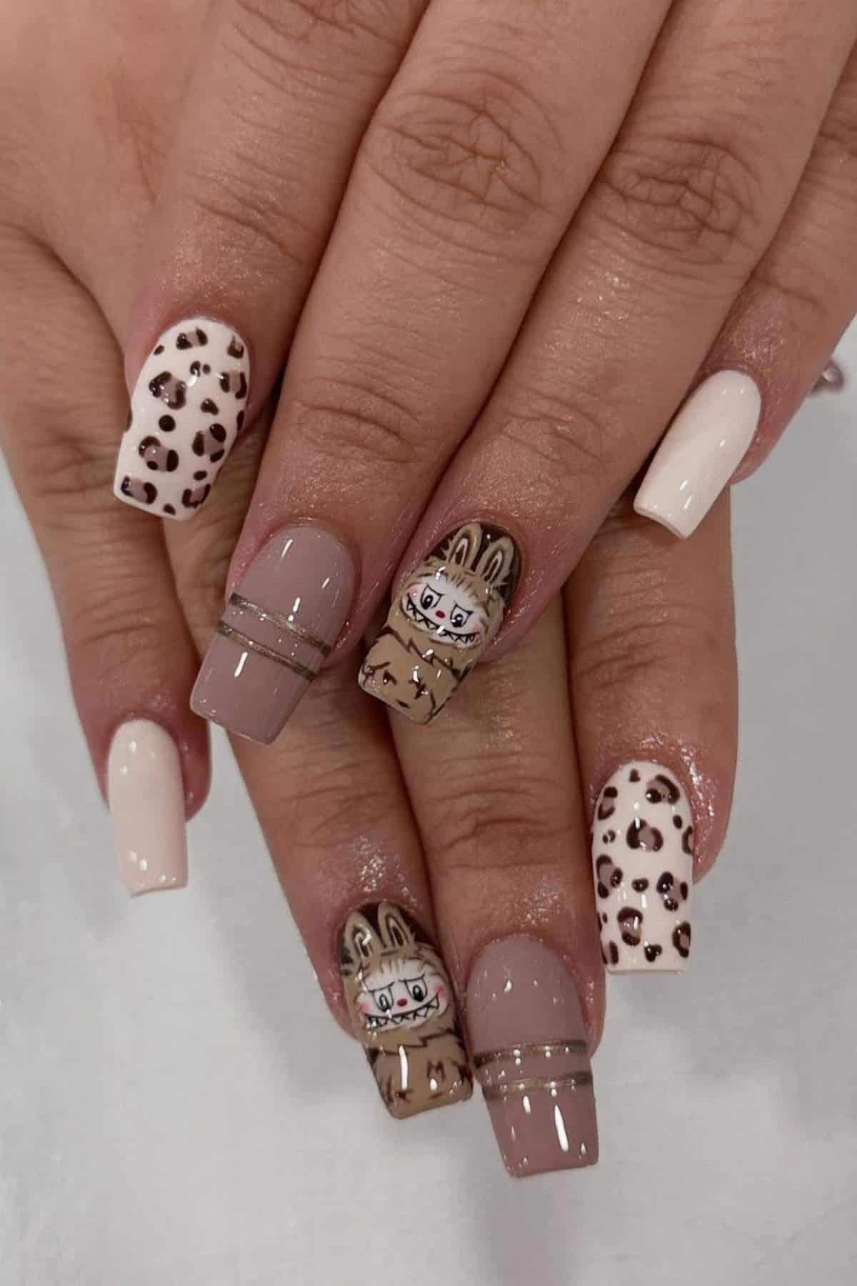 Hands with manicured nails featuring animal print patterns, beige and white nails, and two accent nails with cartoon bunny designs and silver stripes.