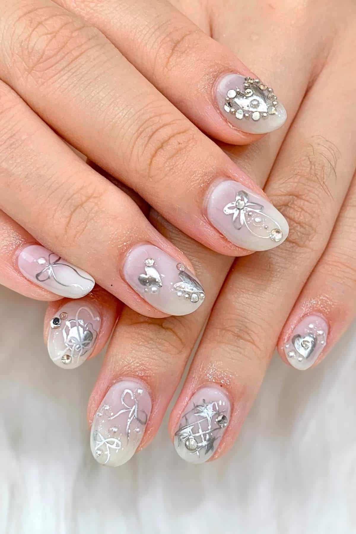 Close-up of hands with short, oval nails featuring a translucent white base and silver 3D nail art designs, including rhinestones, bows, and abstract patterns.