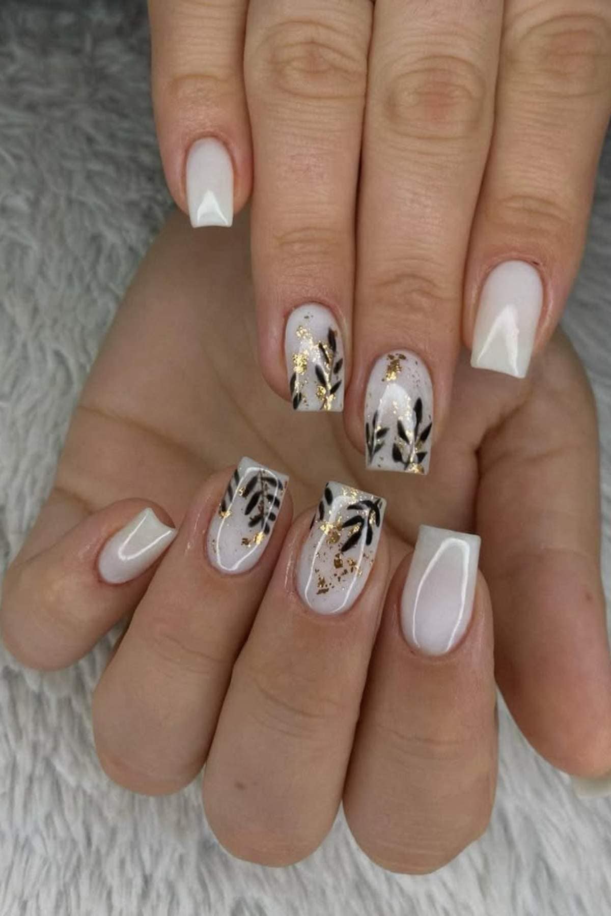 A hand with square-shaped white nails features black leaf designs and gold accents on a few nails, displayed on a textured white surface.