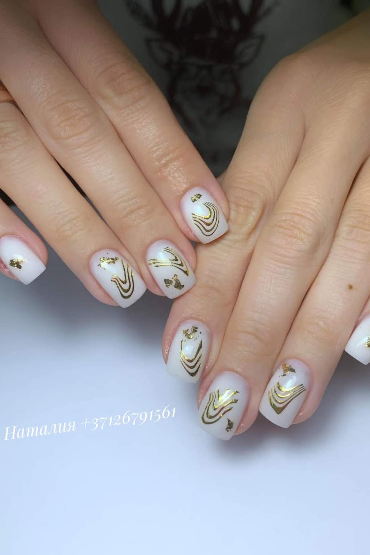 Close-up of hands with short, square-shaped nails featuring white polish and gold decorative lines and flakes.