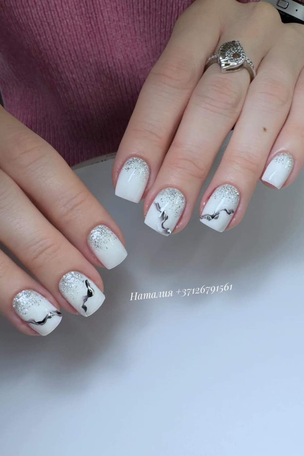 A hand with short, square-shaped nails featuring white polish, silver glitter near the cuticles, and black wavy line designs. The person is wearing a silver ring.