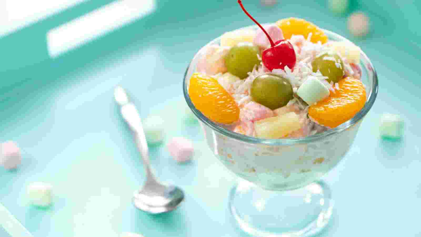 A glass dessert dish filled with fruit salad, topped with grapes, mandarin orange slices, a cherry, and shredded coconut, sits on a light blue surface next to a spoon.