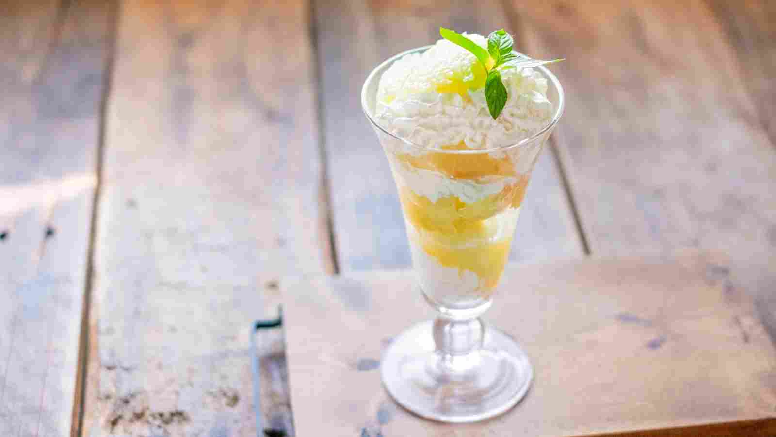 A glass dessert cup filled with layered whipped cream, fruit, and jelly, topped with grated coconut and a sprig of mint, placed on a wooden surface.