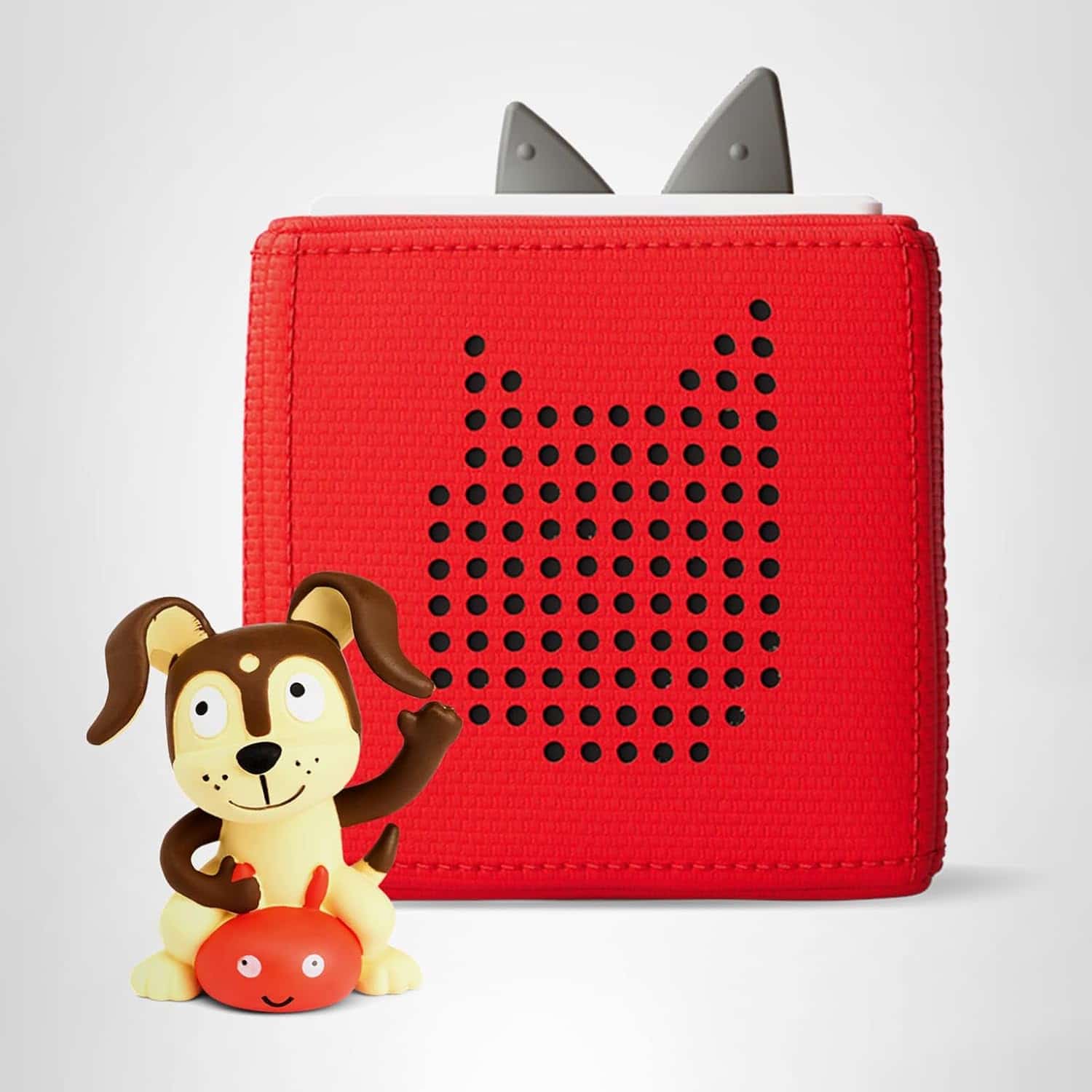 A small plastic dog figurine sits in front of a red, cube-shaped speaker with perforated dots and two grey triangular shapes on top.