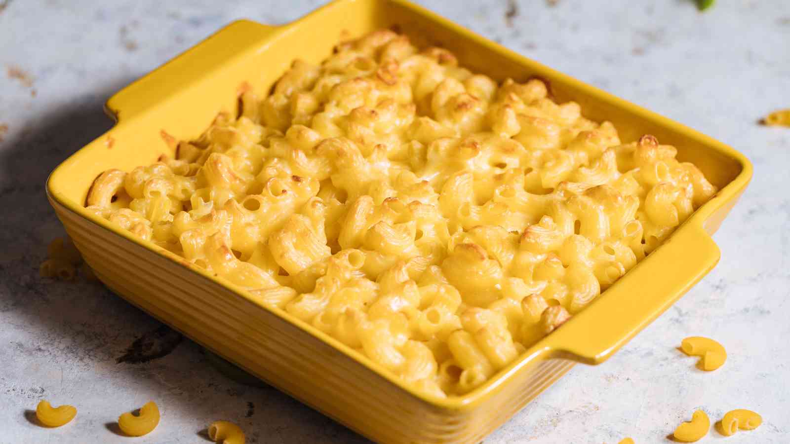 A yellow baking dish filled with baked macaroni and cheese, topped with a light golden brown crust, sits on a light-colored surface.