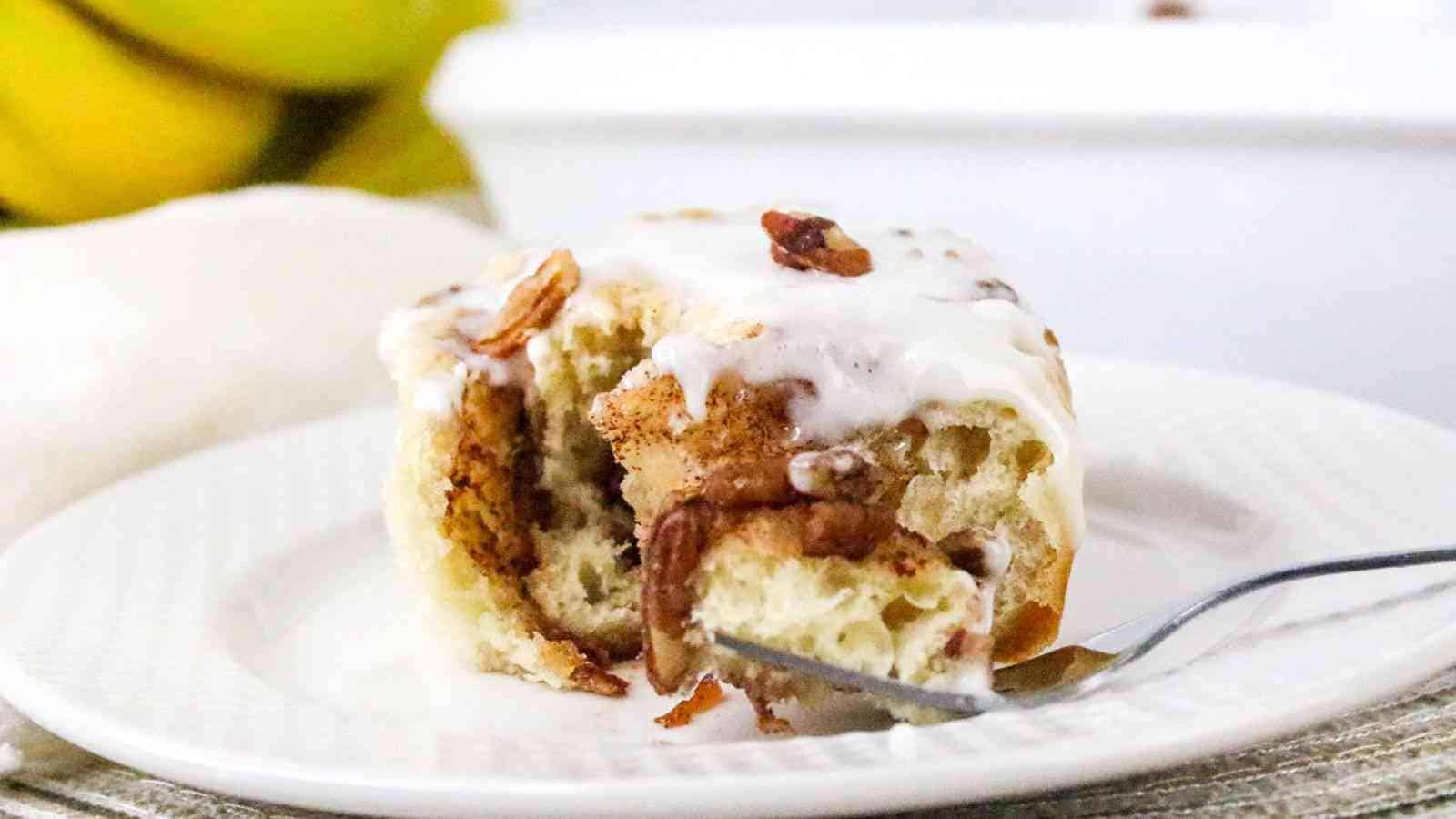 A partially eaten cinnamon roll with icing and pecans on a white plate, with a fork and blurred bananas in the background.