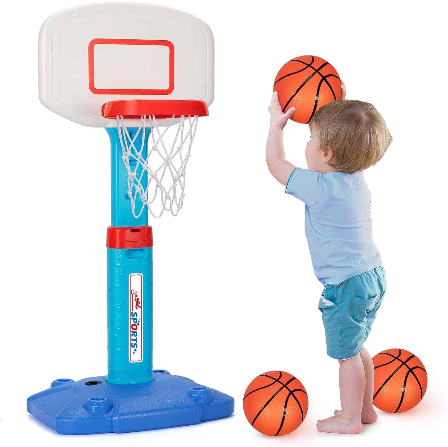 A young child in blue clothes aims to shoot a small basketball into a child-sized indoor basketball hoop, with two more basketballs on the floor nearby.