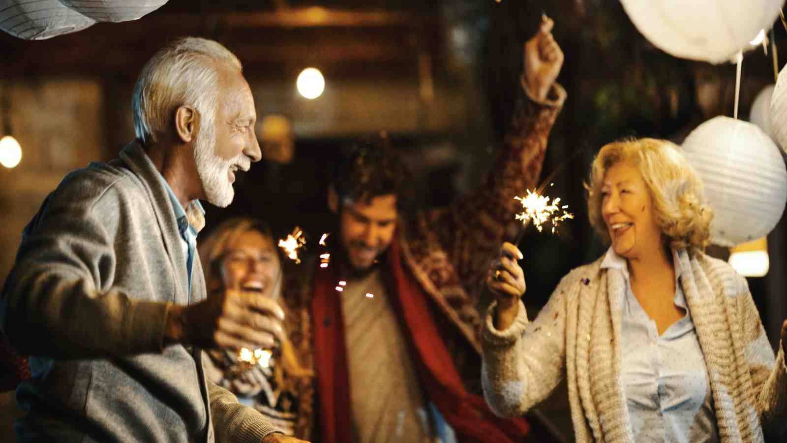 Four adults are smiling and holding sparklers at an outdoor nighttime gathering, surrounded by hanging paper lanterns.