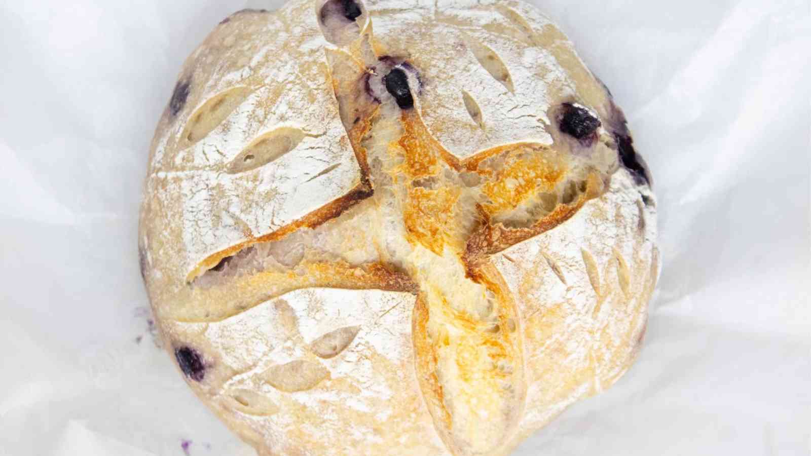 A round loaf of artisan bread with a golden crust, decorative scoring on top, and visible blueberries baked into the dough.