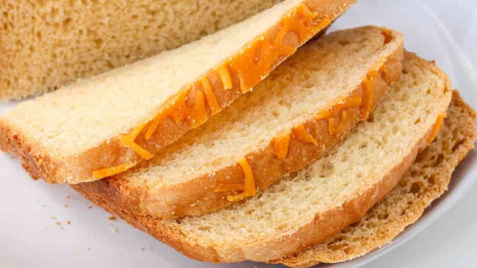 Three slices of bread with visible cheddar cheese pieces are stacked on a plate, with a loaf in the background.