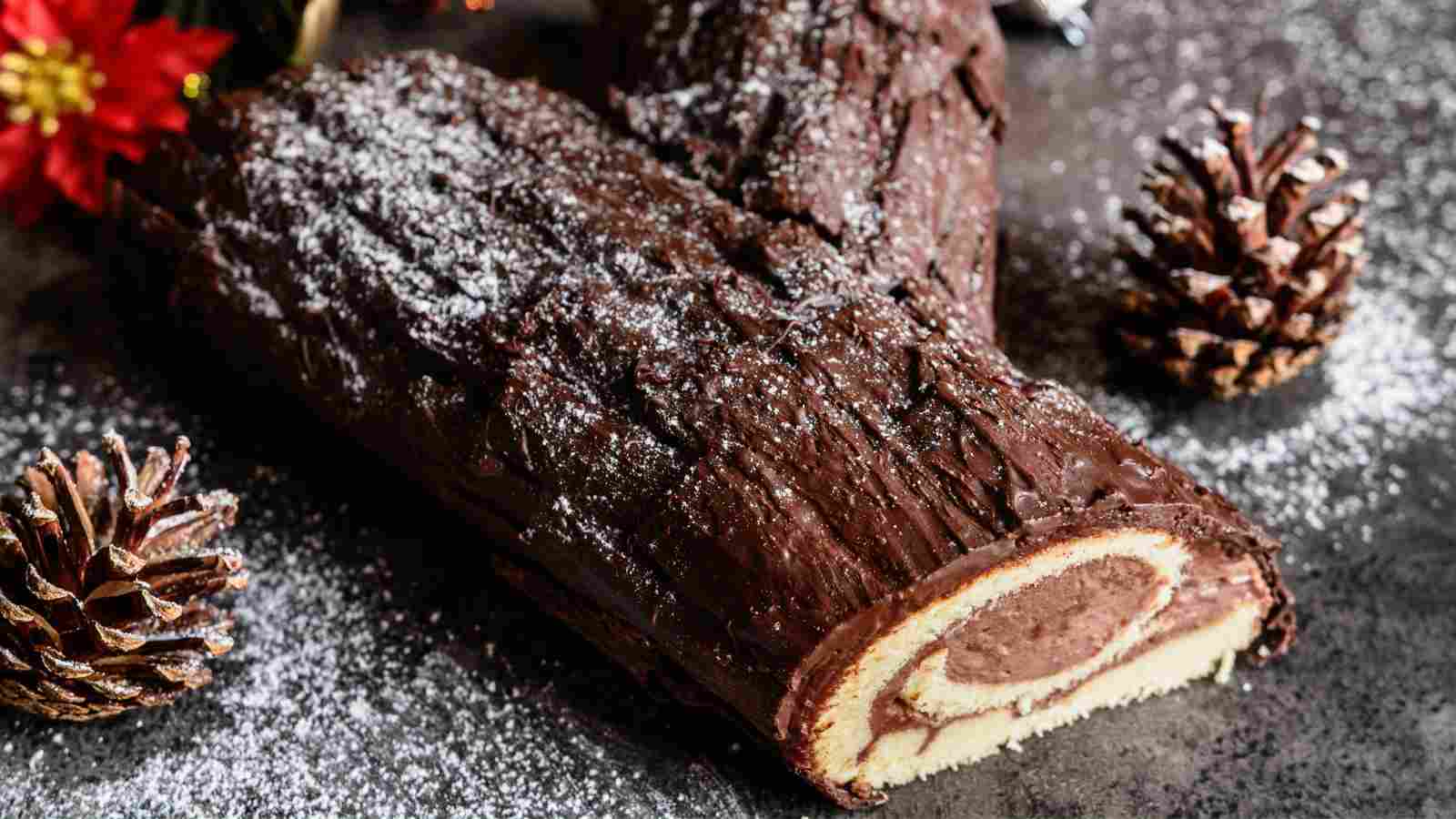 A chocolate yule log cake with a spiral filling is dusted with powdered sugar, surrounded by pine cones and festive decor.