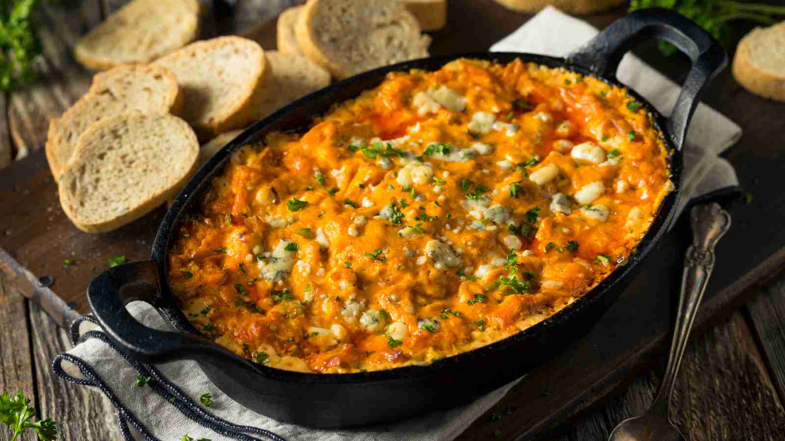 A baked buffalo chicken dip in a black oval dish, topped with melted cheese and herbs, sits on a cloth napkin with sliced bread arranged nearby.