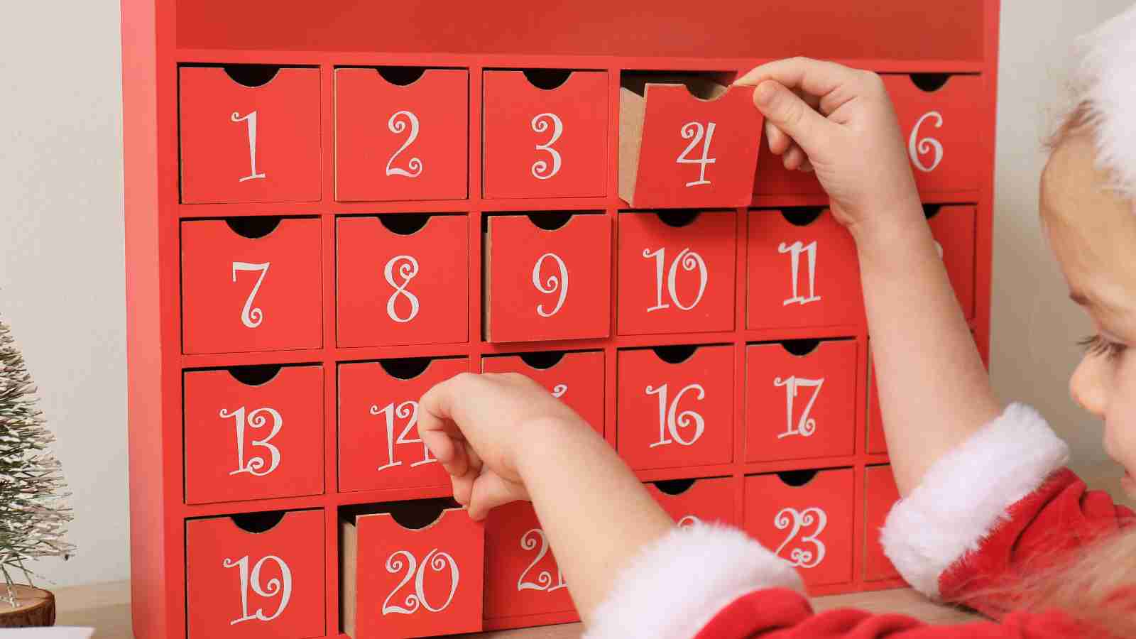 A child opens drawers on a red wooden advent calendar with white numbers.
