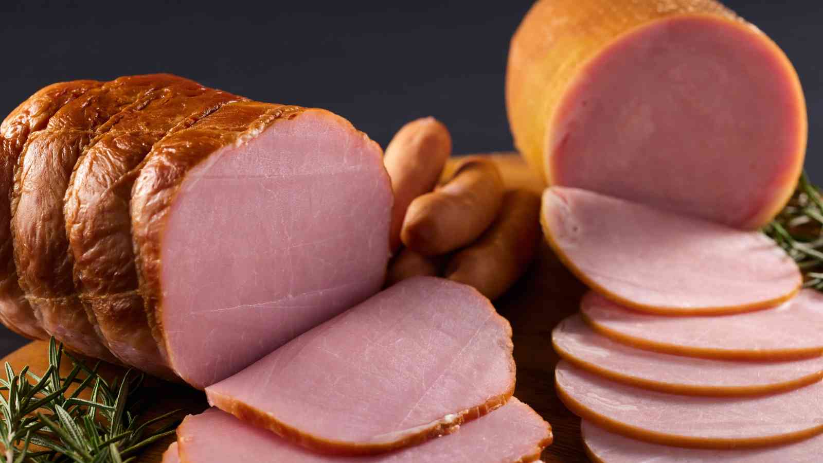 A close-up of sliced ham and sausages on a wooden board, garnished with rosemary sprigs.