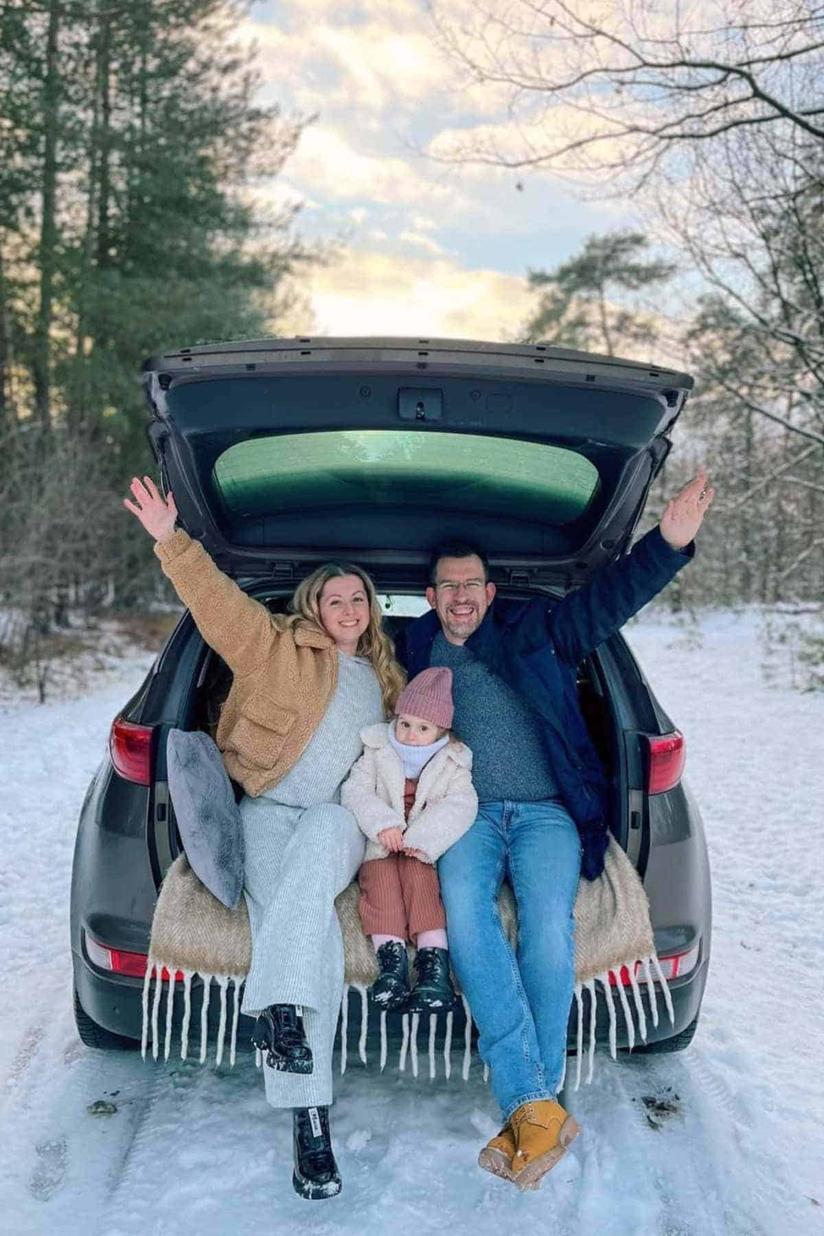 A man, woman, and child sit in the open trunk of a car, smiling with arms raised, surrounded by snow and trees.