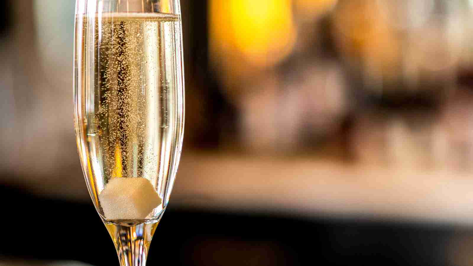 A close-up of a champagne flute with a sugar cube at the bottom and rising bubbles, set against a blurred background.