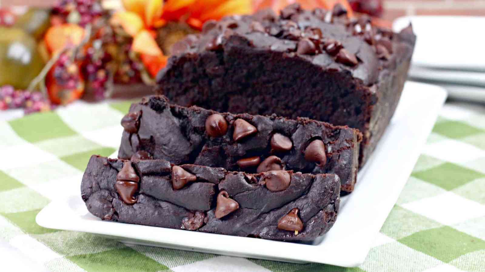 A loaf of chocolate bread topped with chocolate chips, partially sliced on a white rectangular plate, displayed on a green checkered tablecloth.