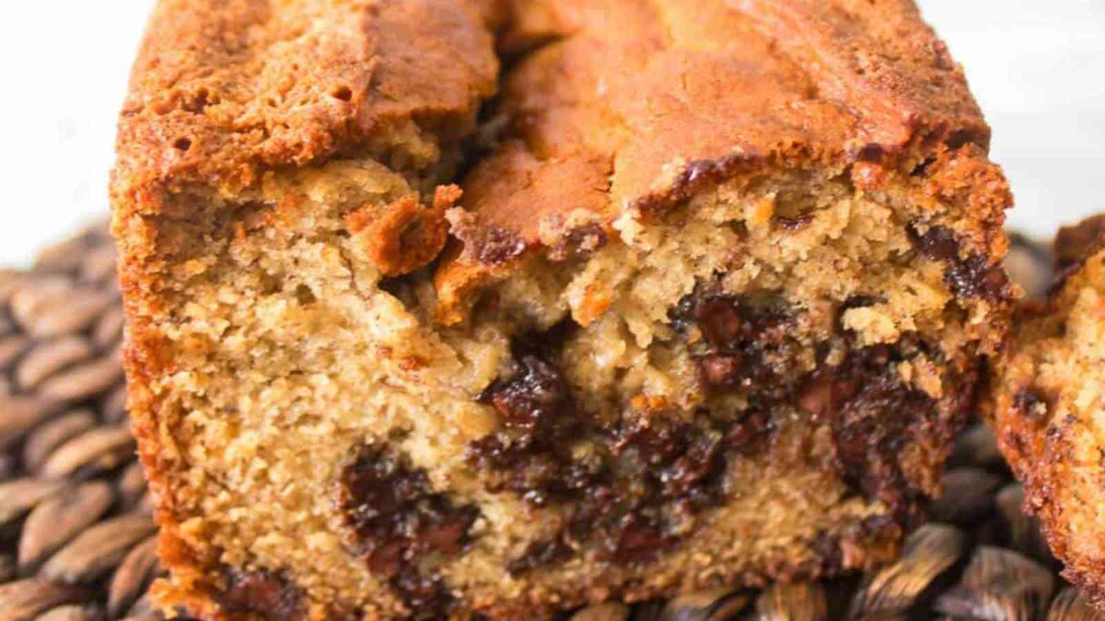 A close-up of a sliced loaf of banana bread with visible chocolate chips on a woven surface.