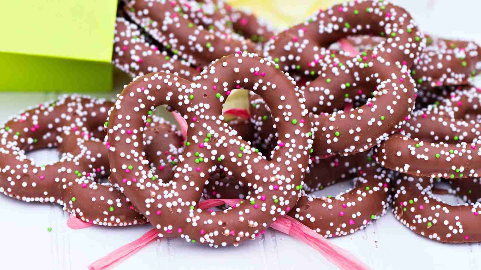 A pile of chocolate-covered pretzels topped with pink, white, and green sprinkles on a light surface.