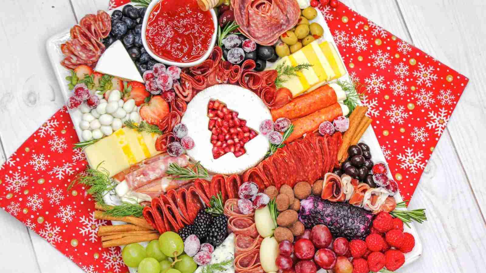 Christmas Charcuterie Board with a large variety of meats, cheeses, fresh fruits and snacks dispayed on a white platter with a red kitchen towel underneath on a white wood board background.