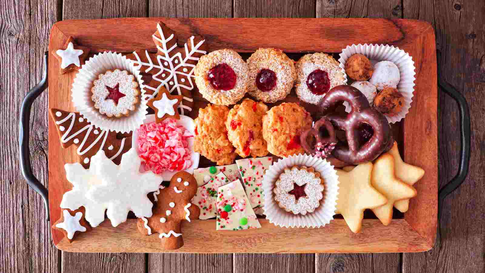 Assorted cookies including gingerbread, snowflake, pretzel, and sandwich cookies with jam are arranged on a wooden tray.