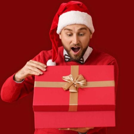 A man in a Santa hat and red sweater looks surprised while opening a large red gift box with a gold ribbon, against a plain red background.
