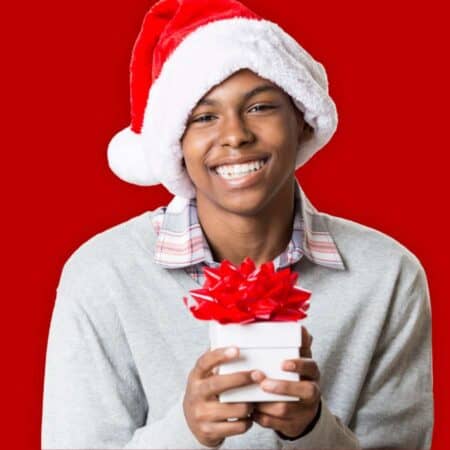 A person wearing a Santa hat and gray sweater holds a white gift box with a red bow, smiling against a solid red background.