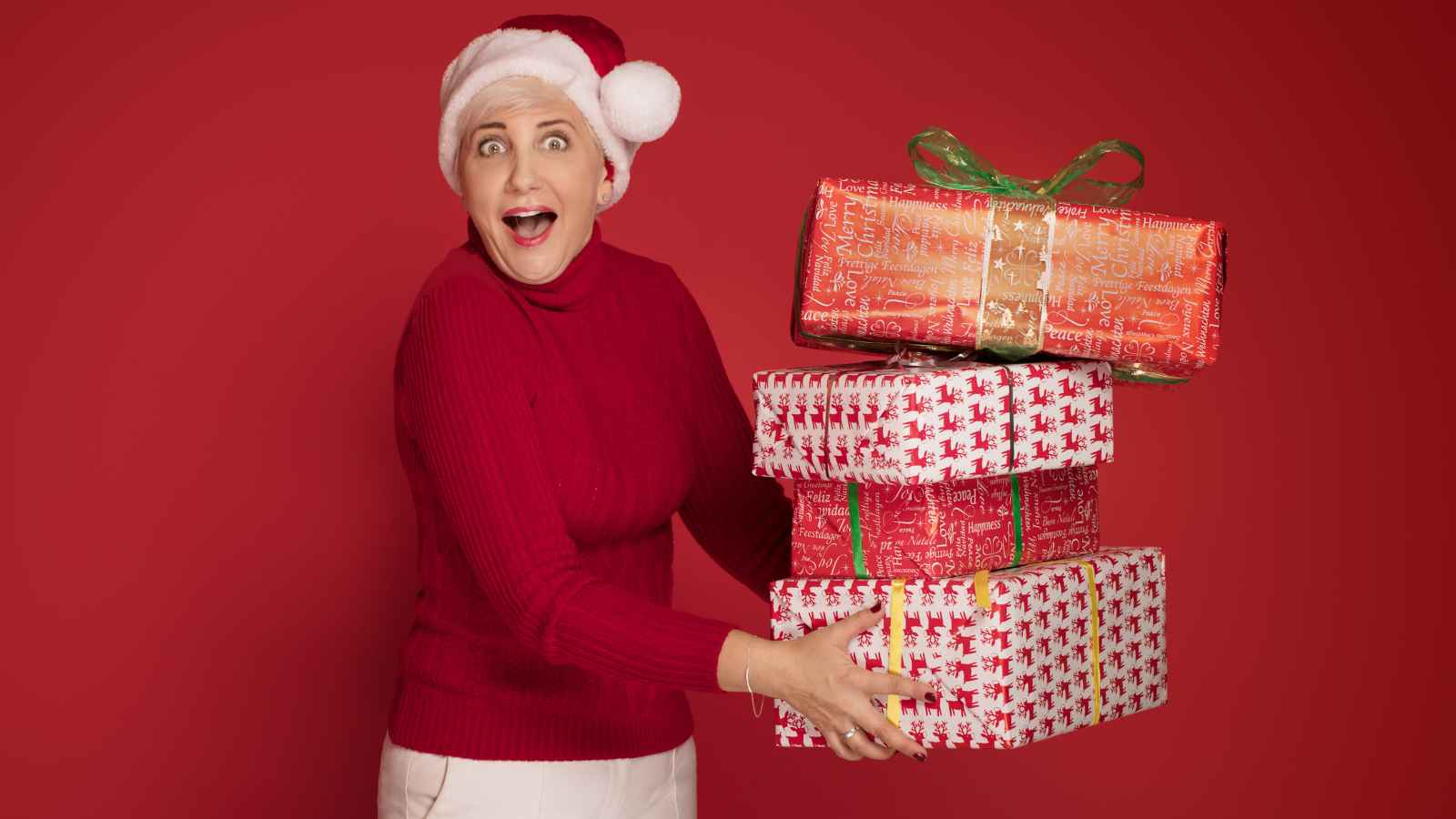 Person in a red sweater and Santa hat holding a stack of three wrapped gifts with a surprised expression against a red background.
