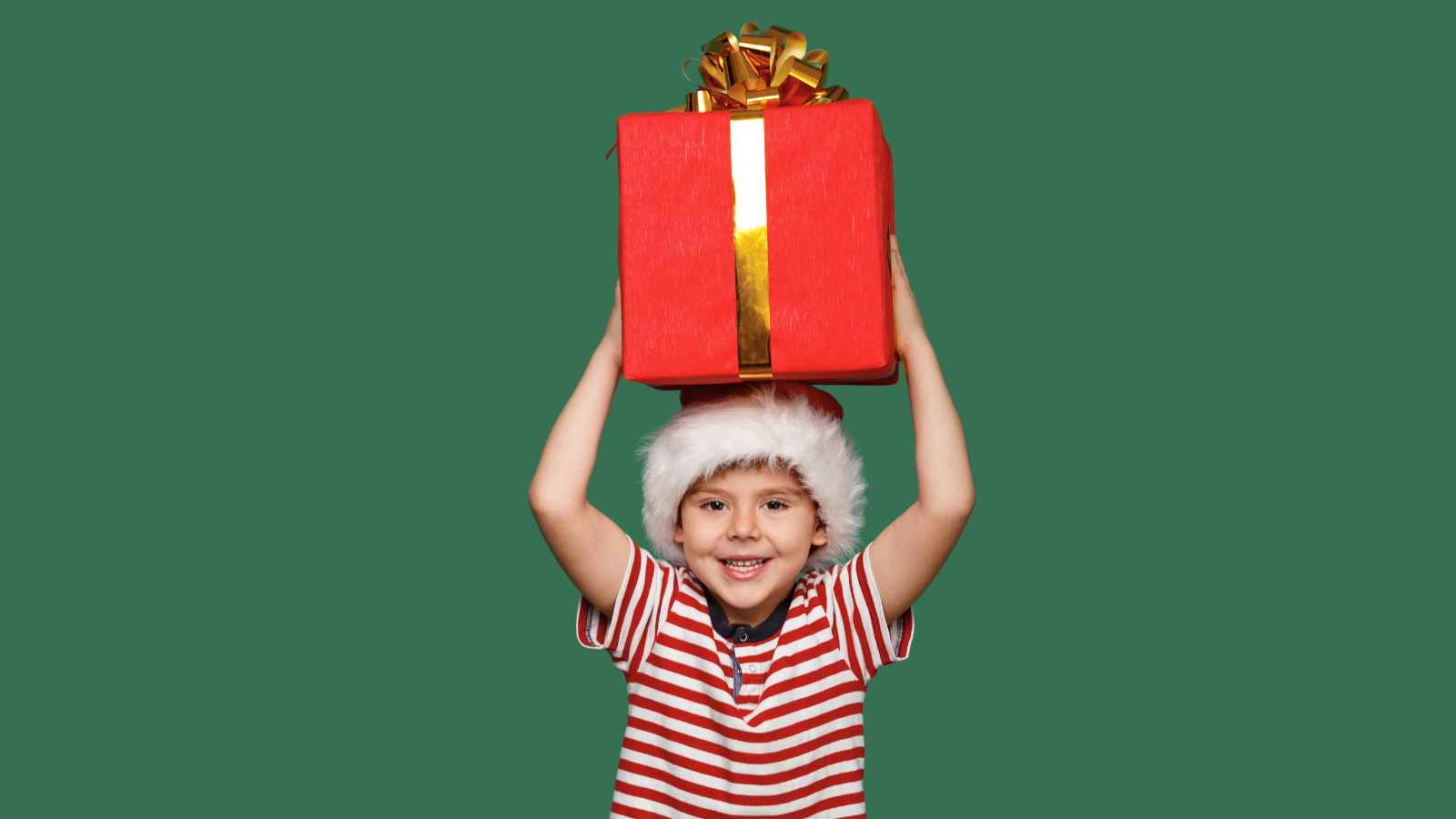 Child in a Santa hat and striped shirt holds a large red gift box with a gold ribbon above their head, standing against a plain green background.