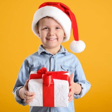 A young child wearing a Santa hat and denim shirt smiles while holding a gift wrapped in white paper with a red ribbon, standing in front of a plain yellow background.