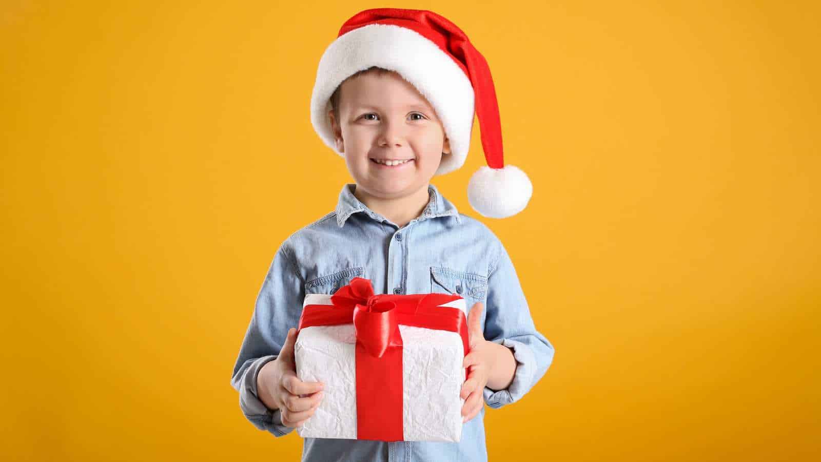 A young child wearing a Santa hat and denim shirt smiles while holding a gift wrapped in white paper with a red ribbon, standing in front of a plain yellow background.