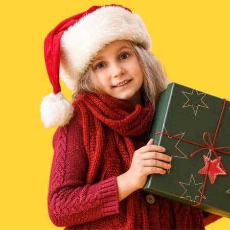 A young girl in a Santa hat and red scarf holds a green gift box with star patterns and a red ornament, standing against a yellow background.