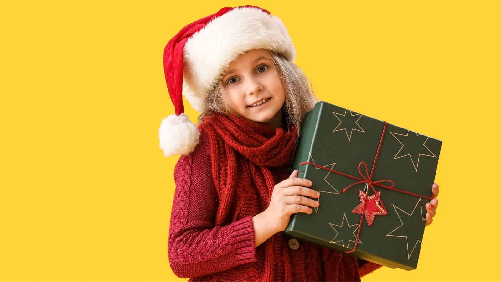 A young girl in a Santa hat and red scarf holds a green gift box with star patterns and a red ornament, standing against a yellow background.