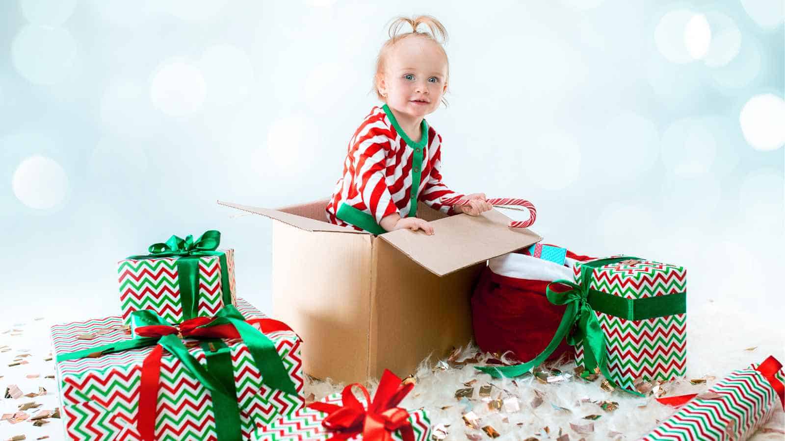 A young child in red and white striped pajamas sits inside a cardboard box, surrounded by wrapped Christmas presents with green and red ribbons.