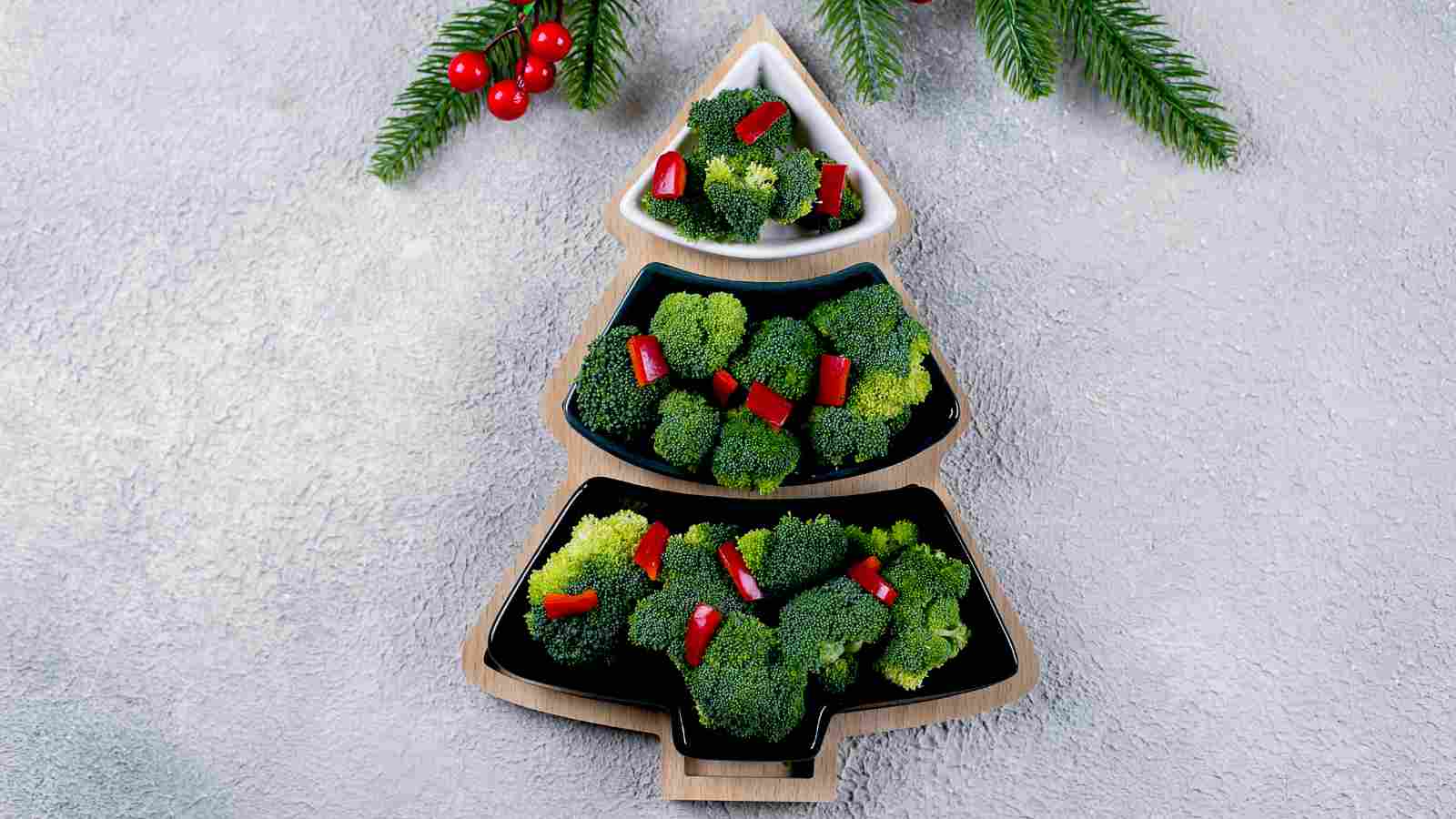 Three tree-shaped trays filled with broccoli florets and red bell pepper pieces, decorated to resemble a Christmas tree, with pine branches and berries nearby.